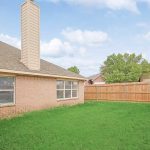 A fenced backyard with green grass, a small tree, and part of a brick house visible, featuring large windows and a chimney. The sky is partly cloudy.
