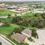 Aerial view of a suburban neighborhood with houses, green lawns, a cul-de-sac, dense trees, and nearby roads. Commercial buildings and parking lots are visible in the background.