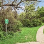 A paved walkway curves through a grassy park area with dense green trees and bushes. A green sign reads "Hiking Trail." A lamppost stands along the path under a partly cloudy sky.