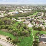 Aerial view of a suburban neighborhood with houses, curved streets, and green spaces bordered by clusters of trees. Roads and distant buildings are visible in the background under a cloudy sky.