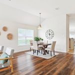 A bright, open dining area with a wooden table, six upholstered chairs, wall clock, and decorative wall baskets. Two armchairs with green cushions sit on a wooden floor near large windows and a view into the kitchen.