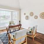Bright home office with a wooden desk facing a large bay window, two cushioned chairs, a black rug, hardwood floors, and decorative wicker wall hangings on a textured white wall.
