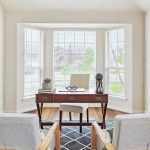 Bright home office with a wooden desk facing a large bay window, two light gray chairs, indoor plants, decorative items on the desk, and three round woven wall hangings on the right wall.