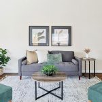 A modern living room with a gray sofa, green armchairs, a round wooden coffee table, and decorative plants. Two framed art pieces hang on the wall above the sofa, and a white rug covers the wooden floor.