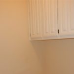 White upper cabinets with vertical grooves are mounted on a plain beige wall in a small, empty room. The lower half of the image shows open space beneath the cabinets.