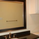 A bathroom with a large framed mirror above a black granite countertop and sink, a towel bar on the wall, a toilet, tiled shower area, and a window reflected in the mirror.