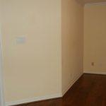 A mostly empty room with beige walls, brown wooden flooring, and a white baseboard. An open doorway on the left reveals a bathroom with a black countertop, white cabinets, and a mirror. A ceiling light is partially visible.