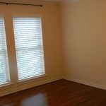 Empty room with light beige walls, two large windows with white blinds, wooden floor, and electrical outlets on the right wall. Natural light comes through the windows.