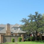 A two-story suburban house with a brick fence, green lawn, and several large trees under a clear blue sky.