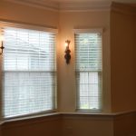 A bright, empty room with light yellow walls, three windows with white blinds, two decorative wall sconces, and crown molding along the ceiling. Natural light streams in through the windows.