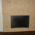 A beige tiled fireplace with a black frame is centered between a glass door with blinds on the left and a window with horizontal blinds on the right, above a dark wood floor.