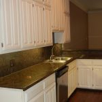 A modern kitchen with white cabinets, dark granite countertops, a stainless steel sink, dishwasher, and wood flooring. There is a door with blinds at the back and under-cabinet lighting brightening the workspace.