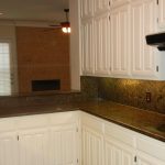 A modern kitchen with white cabinets, dark granite countertops and backsplash, a double sink, and a stove. The kitchen opens up to a living area with a ceiling fan, fireplace, and a glass door to the outside.