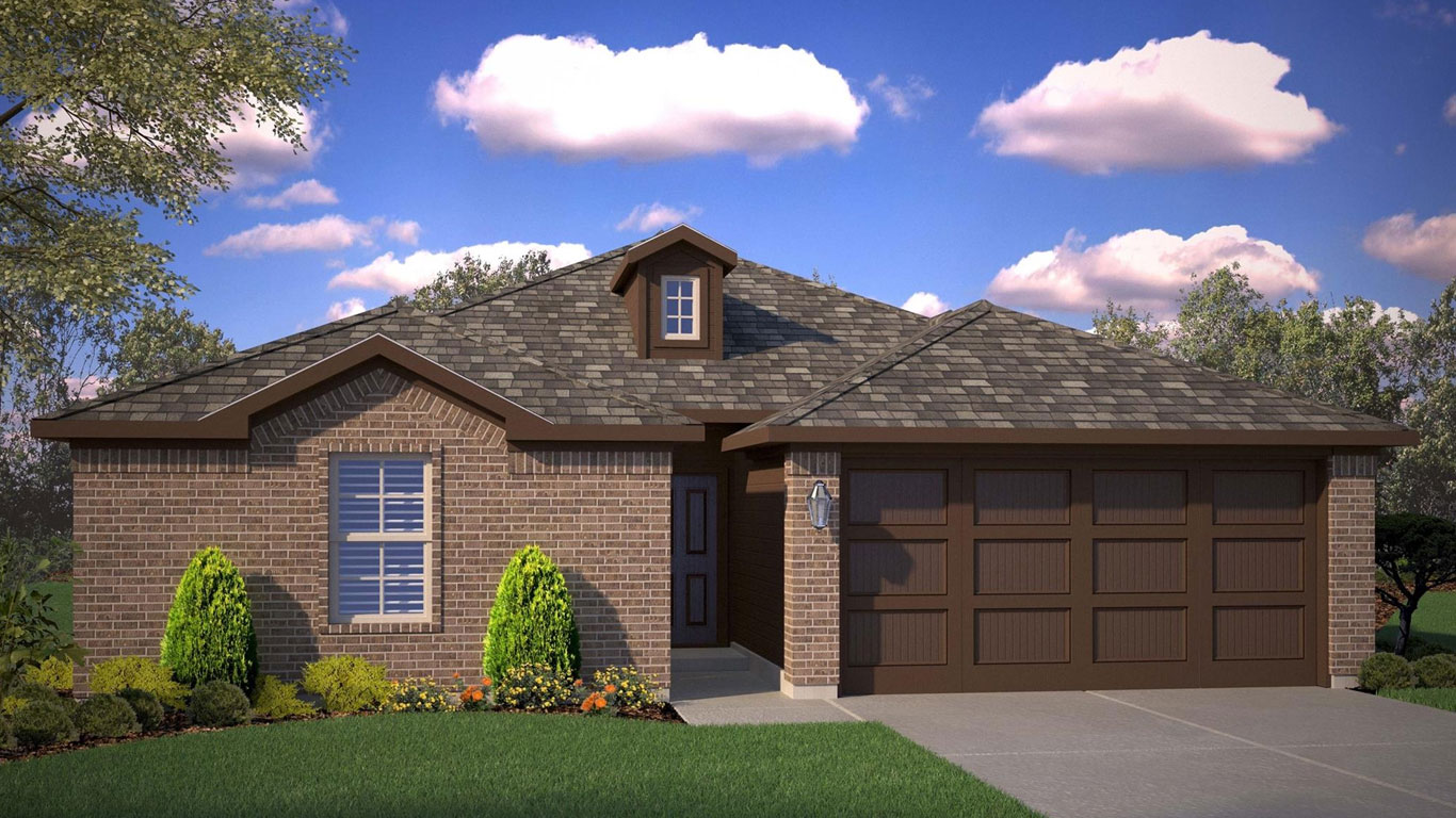 A single-story brick house with a gabled roof, a small dormer, two-car garage, and neatly trimmed bushes and flowers in the front yard under a blue sky with scattered clouds.