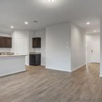Open concept kitchen and living area with wood flooring, dark wood cabinets, stainless steel appliances, granite countertops, and neutral-colored walls. A hallway leads to the front door. Room is unfurnished.