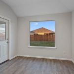 A small, empty room with light gray walls, a white door with a window, wood-look flooring, and a large window showing a fenced backyard and neighboring houses.