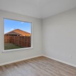 A small empty room with light-colored walls, wood-style flooring, and a large window showing a fenced backyard and part of a neighboring house under a blue sky.