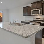 Modern kitchen with dark wood cabinets, stainless steel appliances, a microwave above a gas stove, and a large granite island with a sink. Sunlight streams through a window overlooking a fenced backyard.