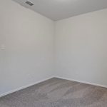 Empty room with light gray walls, a white ceiling, and gray carpeted floor; an open doorway leads to a hallway with wood flooring. Vent and electrical outlets visible on the walls.
