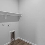 A small laundry room with light gray walls, a wooden floor, a white wall shelf, utility hookups, electrical outlets, and a pet door at the bottom of the wall.