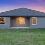 A modern single-story house with gray siding, large windows, and a covered back patio, overlooking a neatly mowed green lawn and enclosed by a wooden fence at sunset.