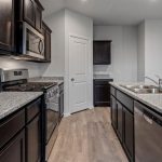 Modern kitchen with dark cabinets, granite countertops, stainless steel appliances, a double sink, dishwasher, gas stove, microwave, and light wood flooring. A closed white door and additional cabinets are in the background.