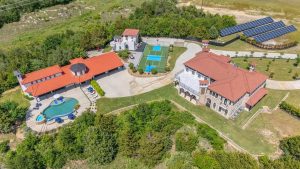 Aerial view of a large estate with a mansion, swimming pool, tennis court, guest house, landscaped grounds, and a solar panel array, surrounded by greenery and open land.