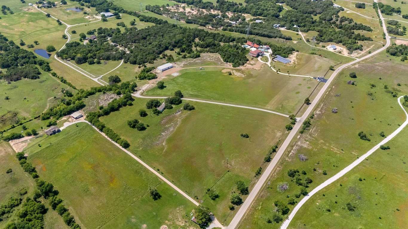 Aerial view of a rural landscape with green fields, scattered trees, winding roads, and a few houses and buildings spread throughout the area.