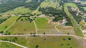Aerial view of a rural landscape with green fields, scattered trees, winding roads, several small buildings, and fenced areas under a clear sky.