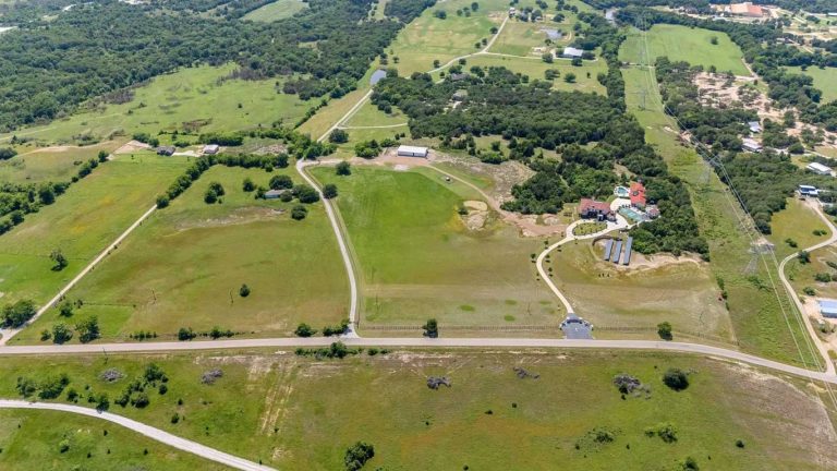 Aerial view of a rural landscape with green fields, scattered trees, winding roads, several small buildings, and fenced areas under a clear sky.