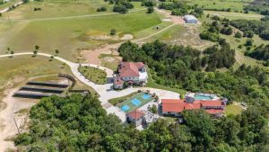 Aerial view of a large estate with two houses, a swimming pool, a tennis court, driveways, and surrounding greenery and open fields.