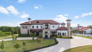A large Mediterranean-style house with a red tile roof, arched windows, and stone exterior, surrounded by manicured lawns, young trees, and a circular driveway under a bright blue sky.