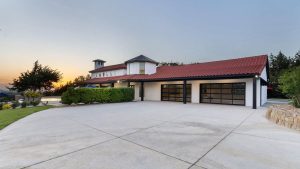 Modern house with red-tiled roof, large driveway, three glass garage doors, and surrounding greenery. The sun is setting in the background, casting a warm glow over the property.