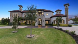 A large, elegant two-story stone house with arched windows, red tile roof, and two tall chimneys, surrounded by a manicured lawn and trees at sunset.