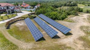Aerial view of three rows of solar panels installed on open land near a large house with a pool, surrounded by trees and greenery.