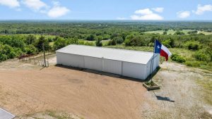 A large white metal building sits on a gravel lot surrounded by trees and greenery. A Texas state flag flies on a tall flagpole near the building under a blue sky with scattered clouds.