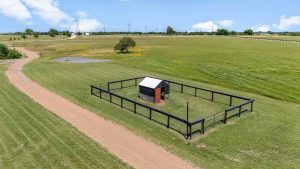 A small fenced enclosure with a shed stands on a grassy field next to a gravel road, surrounded by open landscape, trees, a pond, and distant power lines under a blue sky.