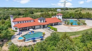 Aerial view of a large villa with a red tile roof, a curved swimming pool, shaded lounge areas, landscaped greenery, a tennis or basketball court, and open fields in the background.