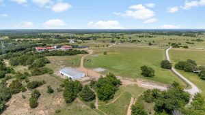Aerial view of a rural landscape with open green fields, scattered trees, a large metal barn, a dirt road, and a few buildings in the distance under a blue sky with scattered clouds.
