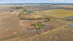 Aerial view of farmland with scattered houses, fields, and a small pond surrounded by trees, under a partly cloudy sky.