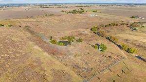 Aerial view of a rural landscape featuring open fields, a small pond surrounded by dry grasses, scattered trees, and a few distant houses under a partly cloudy sky.
