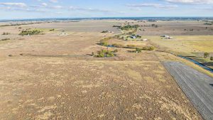 Aerial view of a vast rural landscape with open fields, scattered trees, small ponds, and a few farmhouses under a blue sky with clouds.