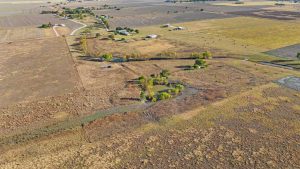 Aerial view of a rural landscape with open fields, scattered trees, small ponds, and a few houses surrounded by farmland under a clear sky.