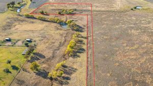 Aerial view of a rural property outlined in red, featuring open fields, clusters of trees, two ponds, and a small creek running through the land. neighboring fields and a few small structures are also visible.