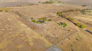 Aerial view of a rural landscape with dry fields, a small pond surrounded by trees, and scattered farm buildings in the distance under a clear sky.