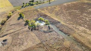 Aerial view of a rural landscape featuring a small pond surrounded by trees, open fields, and patches of dry grass with some farmland in the background.