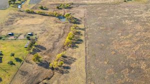 Aerial view of a rural landscape with a small pond, scattered trees, fenced fields, and a cluster of buildings on the left, with open grassy plains on the right.