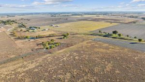 Aerial view of expansive rural farmland with fields, patches of trees, scattered buildings, and a few ponds under a partly cloudy sky. The landscape stretches to the horizon with a mix of green and brown areas.