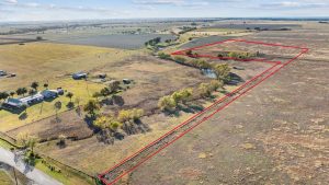 Aerial view of rural farmland with a section outlined in red, bordering a road and extending irregularly across open fields with scattered trees, buildings, and a pond visible nearby.