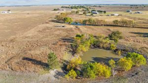 Aerial view of a rural landscape with a small pond surrounded by trees, open fields, and scattered houses in the distance under a partly cloudy sky.